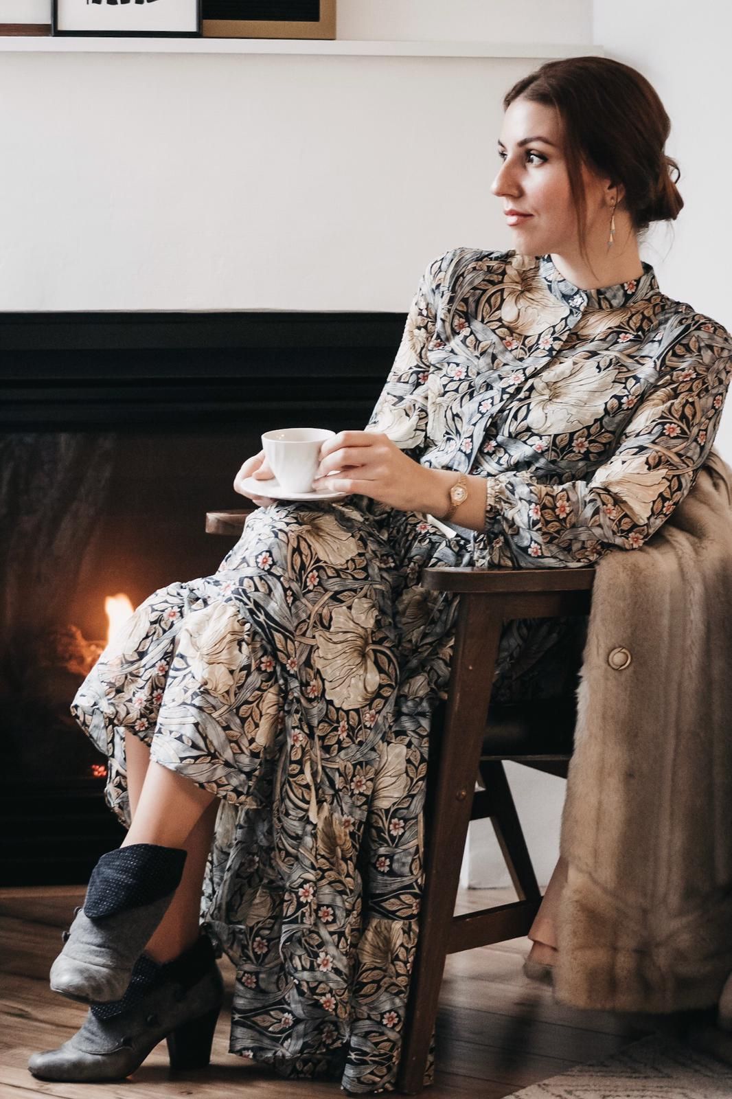 Author of lifestyle blog sitting by fireplace with a cup of coffee wearing a floral dress.